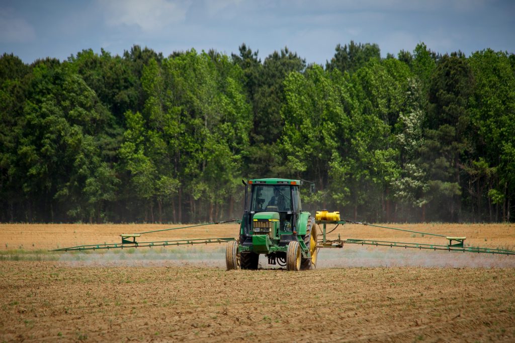 A modern green tractor applying fertiliser with boom sprayer on a dry field surrounded by lush green forest—symbolising modern agriculture and market competition